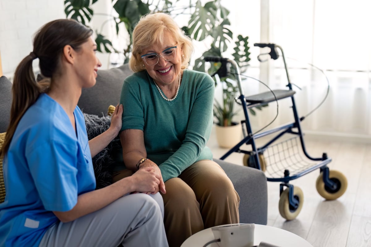 A caregiver and an older woman sitting together in a comfortable living room, holding hands and talking