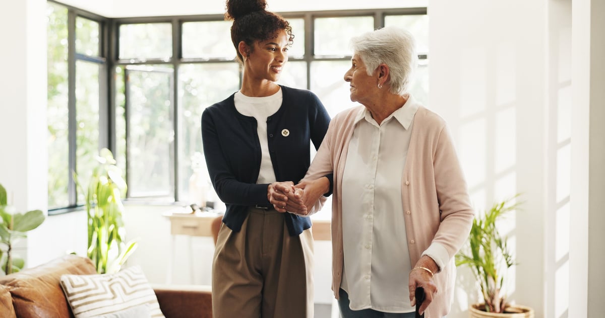 A caregiver holding hands with an older woman as she walks with a cane at home