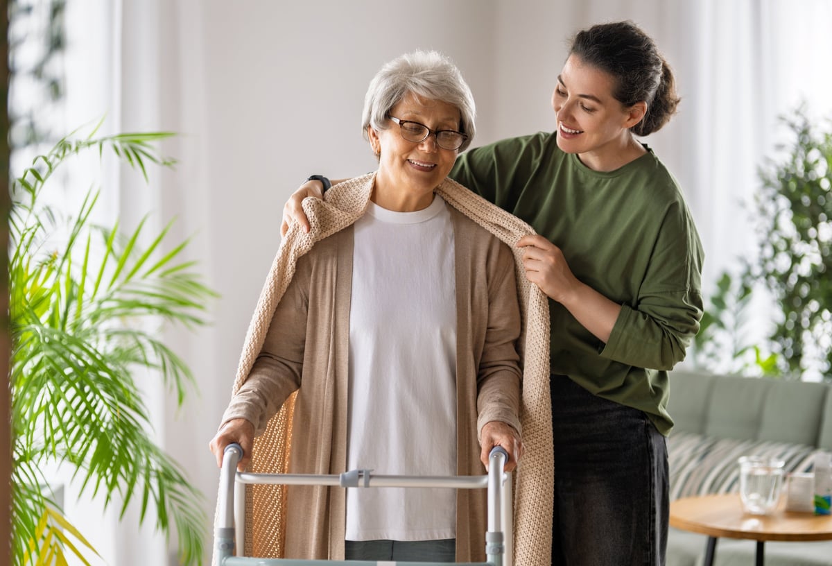 A caregiver gently placing a shawl on an older woman who uses a walker in a bright living room